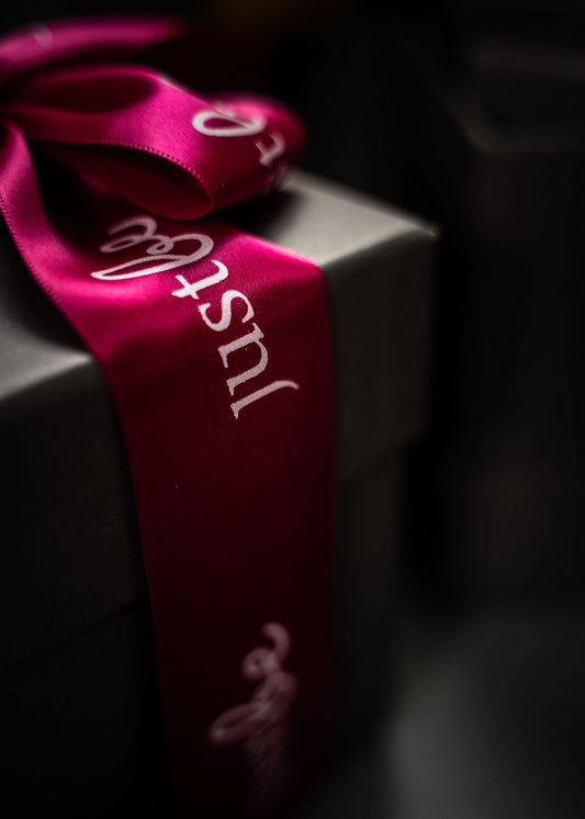 Close-up of a dark gift box tied with a deep pink satin ribbon featuring the JustBe Botanicals logo. The lighting is warm and festive.