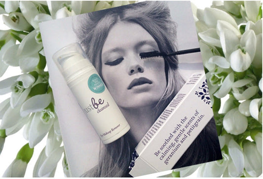 black and white poster of model applying mascara featuring JustBe Botanicals Eye Makeup Remover bottle on a monochrome beauty editorial with snowdrops background. Text on packaging highlights natural calming properties of geranium and petitgrain.