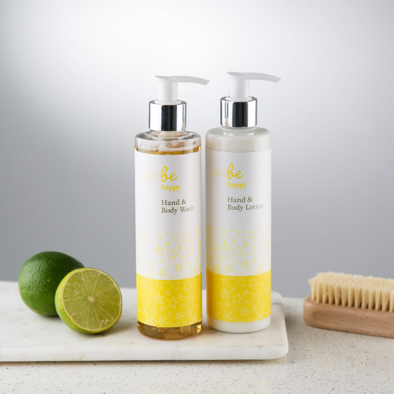 JustBe Happy Hand & Body Wash and Lotion in white bottles with yellow star fish pattern, shown with fresh lime and nail brush on a marble tray.