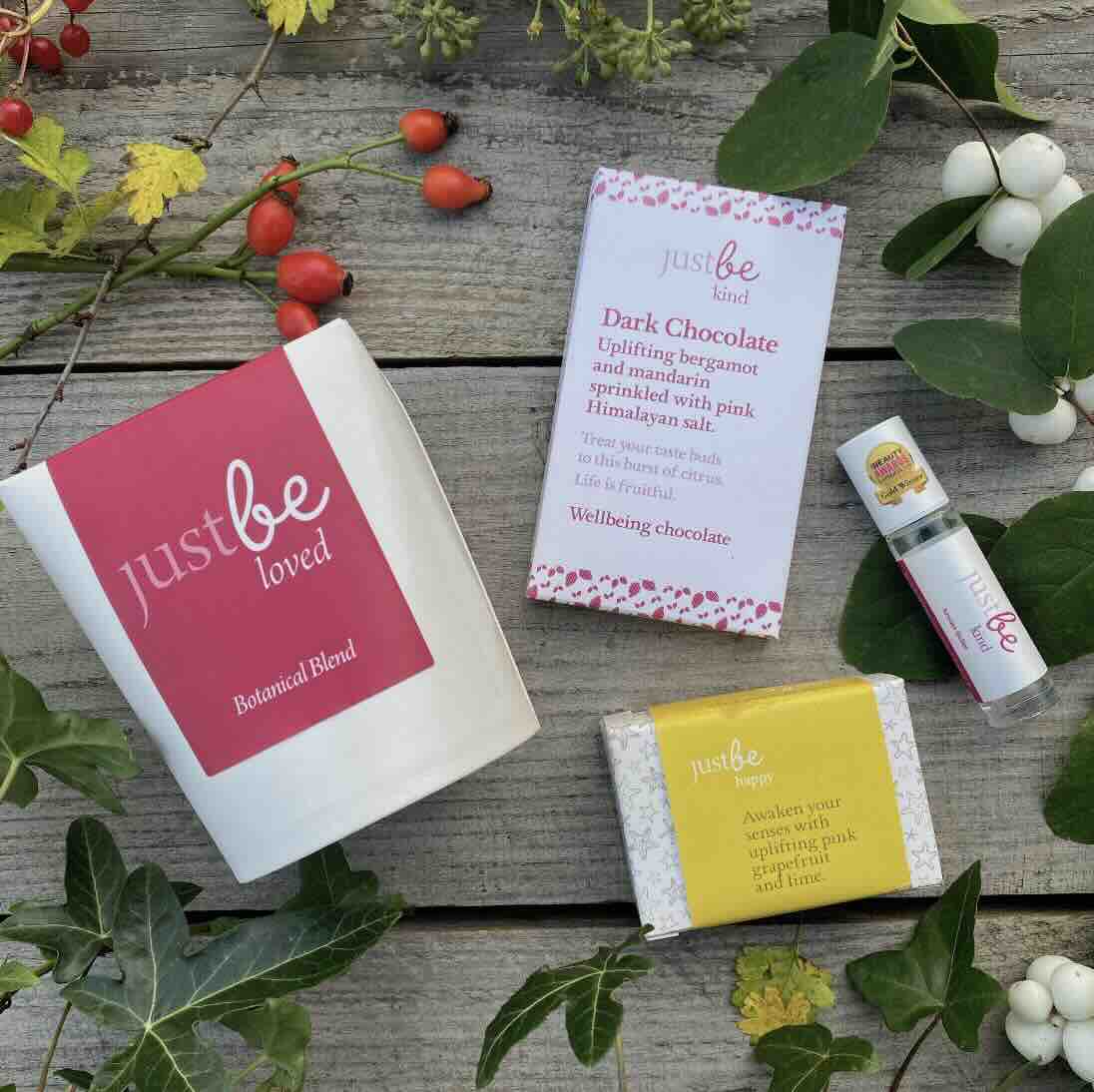JustBe Botanicals wellbeing gift set including Loved herbal tea, Kind dark chocolate bar, Happy soap bar, and Kind aromatherapy rollerball, styled with greenery on rustic wood.