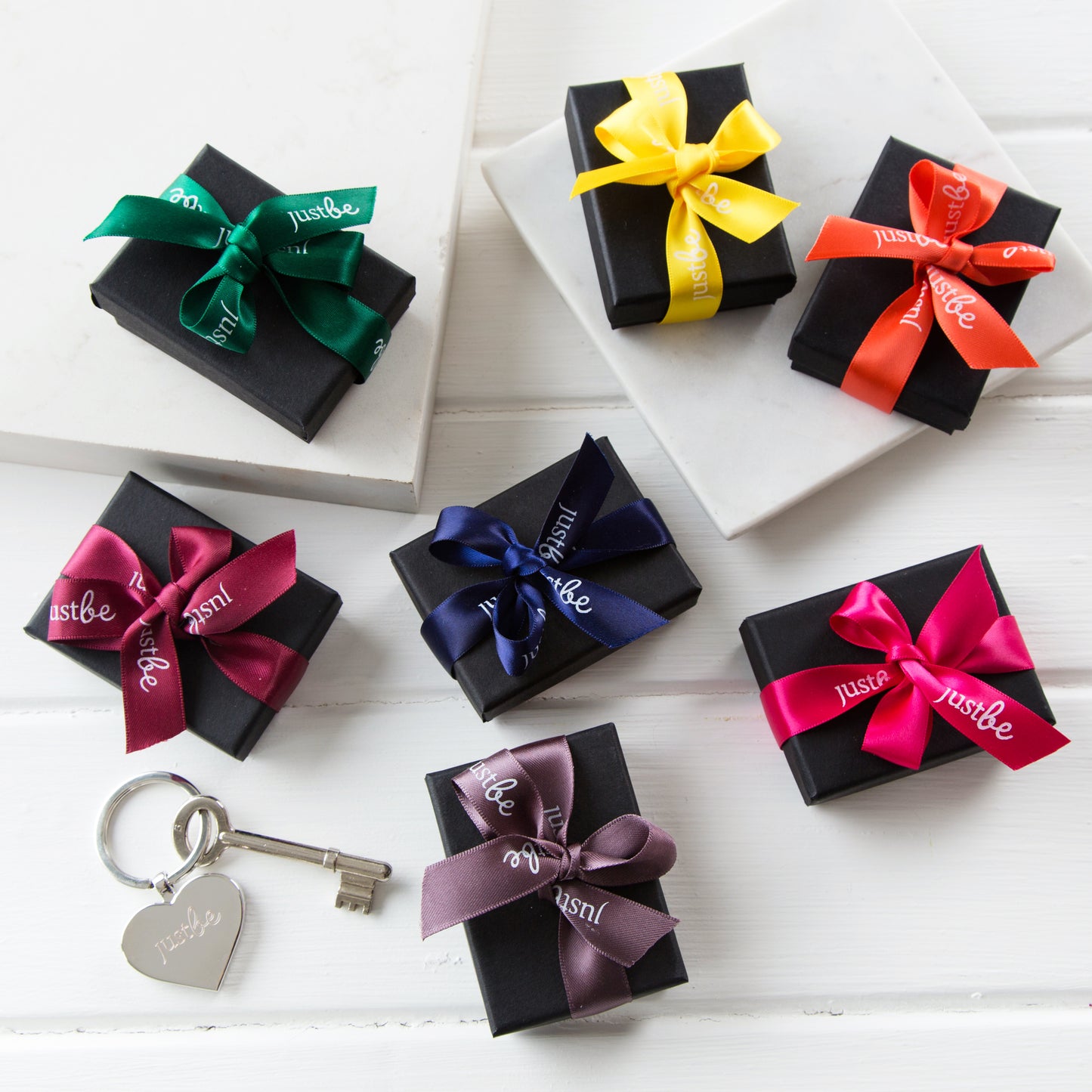 7 small black gift boxes with bright JustBe ribbon sitting beside river heart shaped JustBe engraved keyring