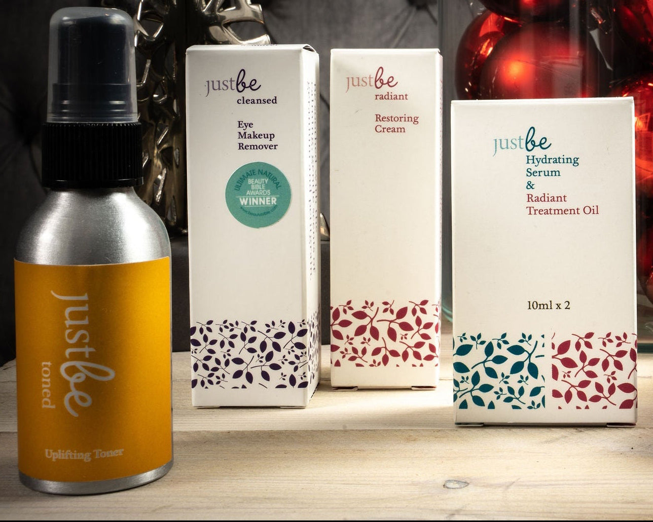 Festive set up of JustBe Uplifting Toern, Eye Makeup Remover, Restoring Cream and Hydrating Serum & Treatment Oil. Pictured in front of river christmas tree decoration and glass container with red Christmas balls
