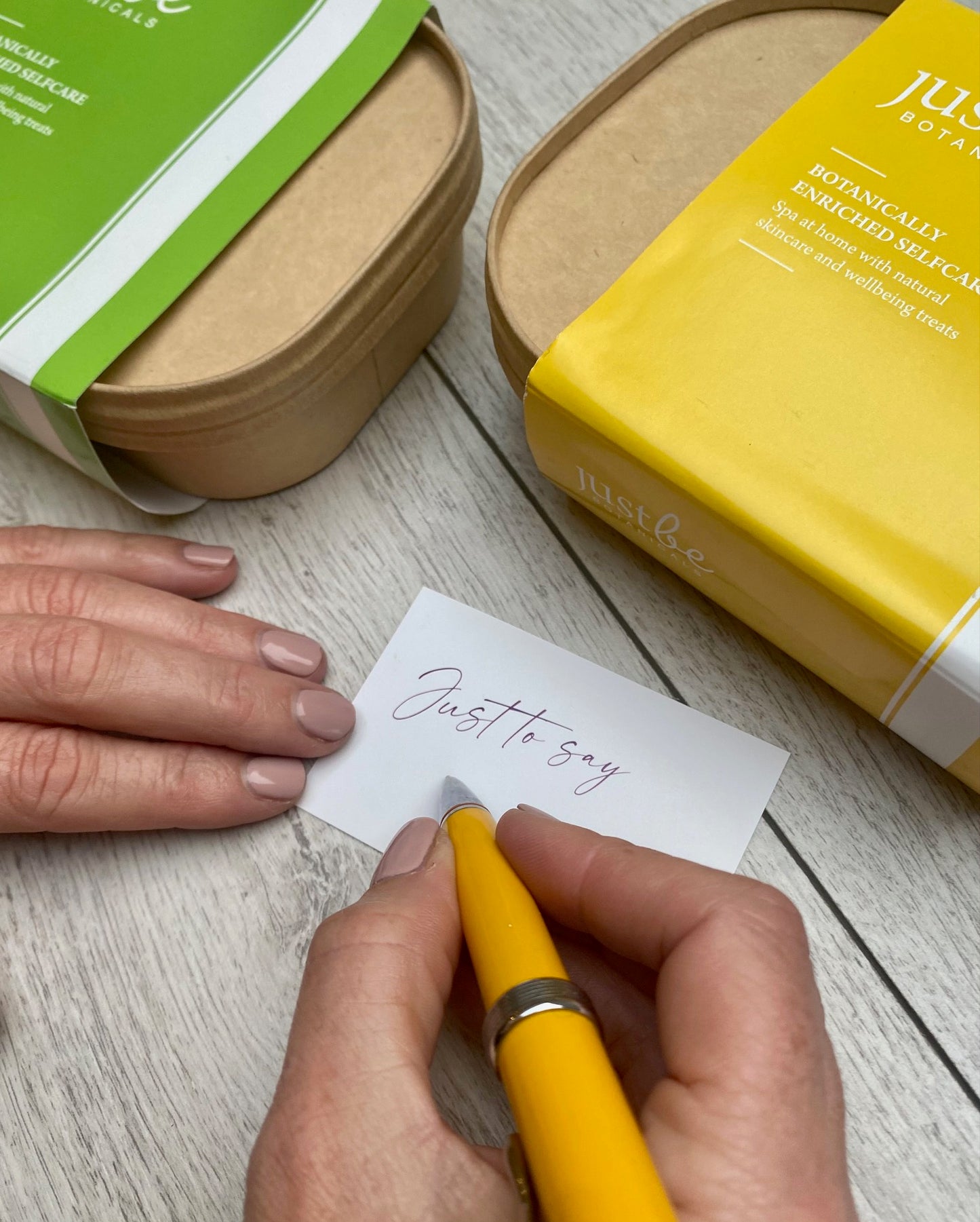 Writing a note on piece of card that says Just to say. Pictured beside JustBe Botanicals green and yellow gift sets.