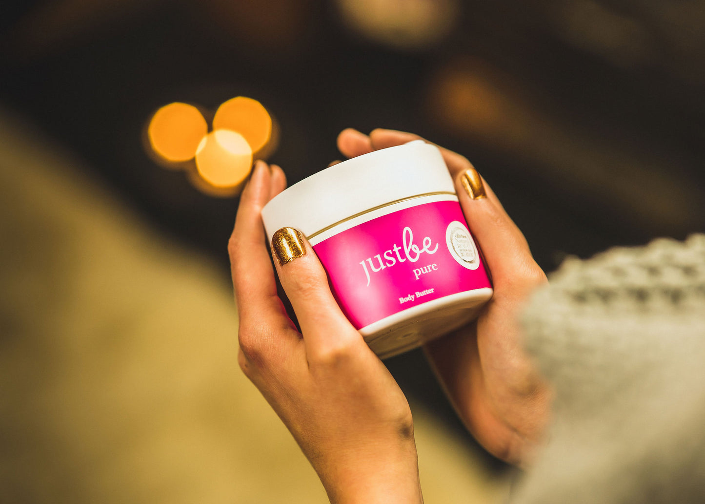 Festive setting of white jar of JustBe Pure Body Butter being held by woman wearing gold nail polish