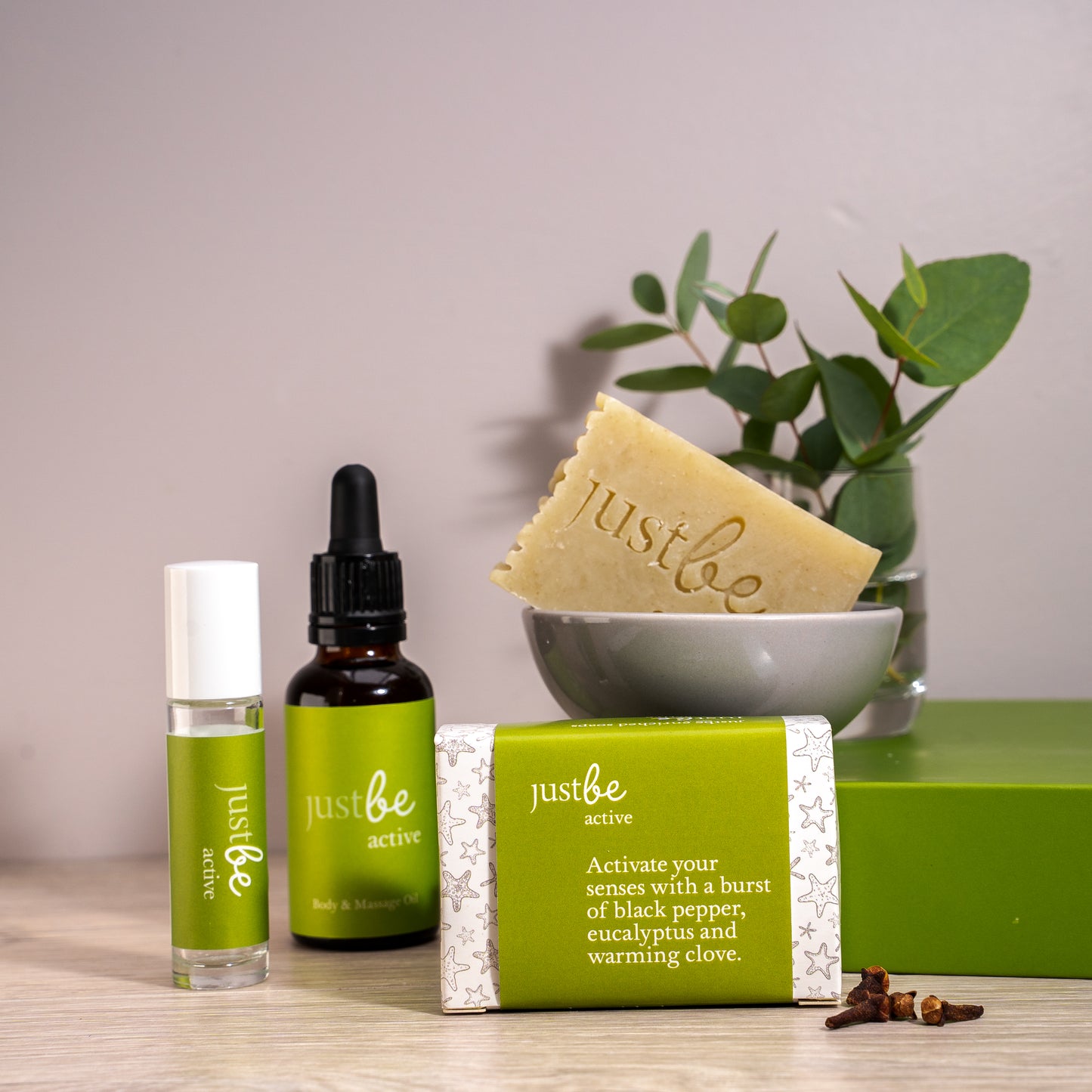 JustBe Active aromatherapy set featuring rollerball, body & massage oil, and natural soap bar all with JustBe green-label displayed with soap bar in a grey dish and eucalyptus leaves and cloves, evoking a fresh and energising vibe.