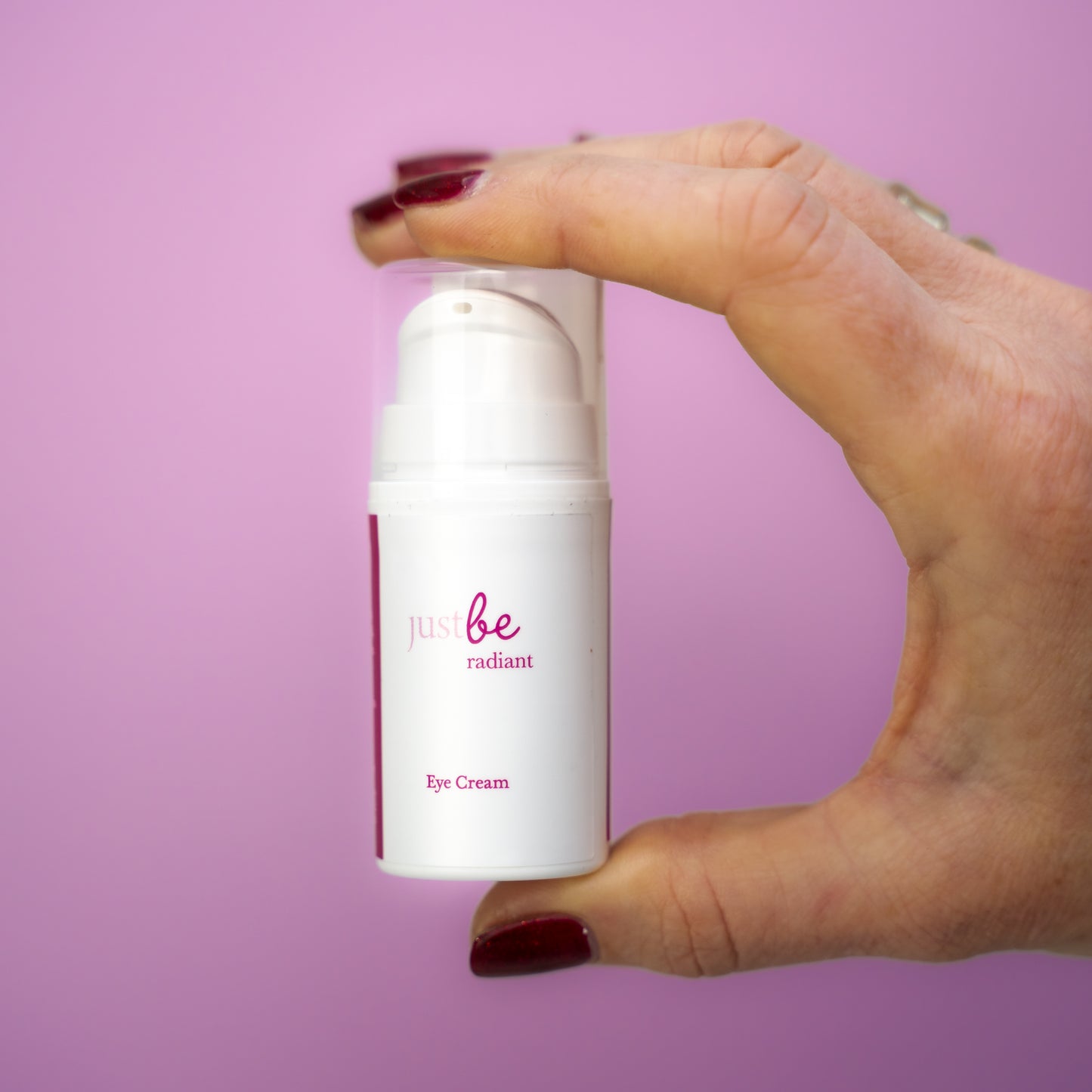 Hand holding JustBe Radiant Eye Cream against a soft pink background, highlighting the compact, travel-friendly packaging