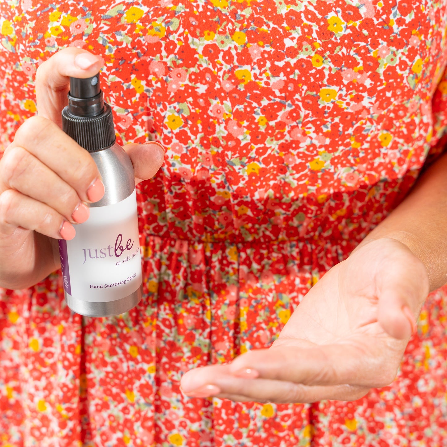 Person spraying JustBe Botanicals "In Safe Hands" hand sanitising spritz into palm, wearing a vibrant red floral dress.