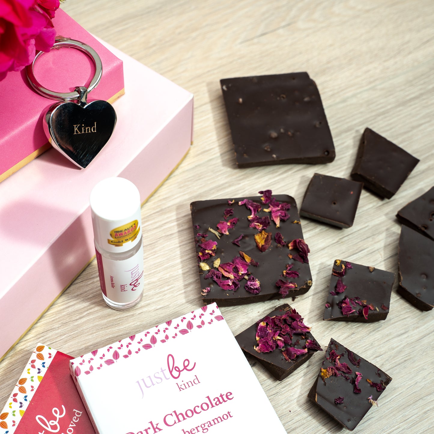 Flatlay of a wellbeing gift set including a heart-shaped keyring engraved with the word kind, an aromatherapy rollerball, and dark chocolate pieces with rose petals beside two JustBe chocolate bars