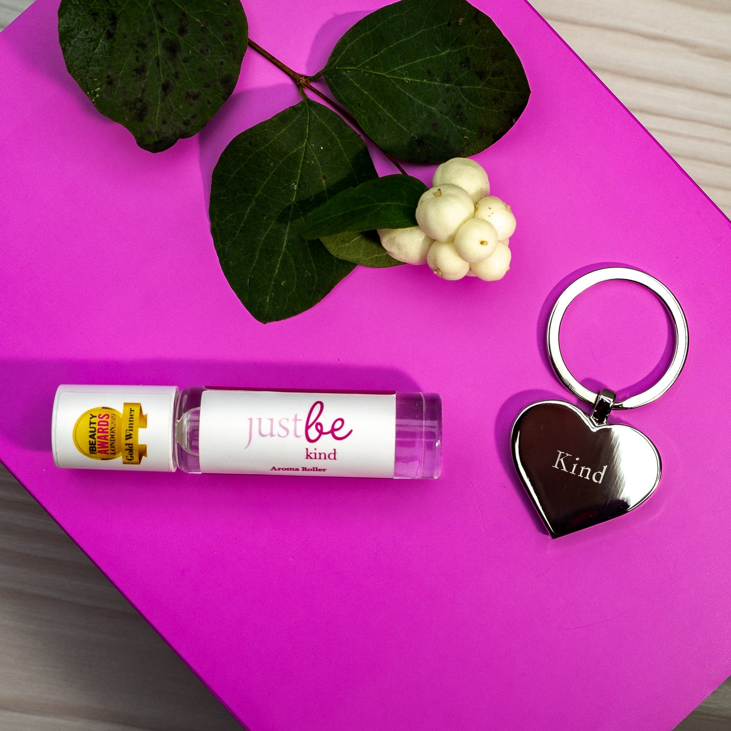 JustBe Kind aromatherapy rollerball and silver heart-shaped keyringengraved with word 'Kind' displayed on a bright pink background with white berries and green leaves.