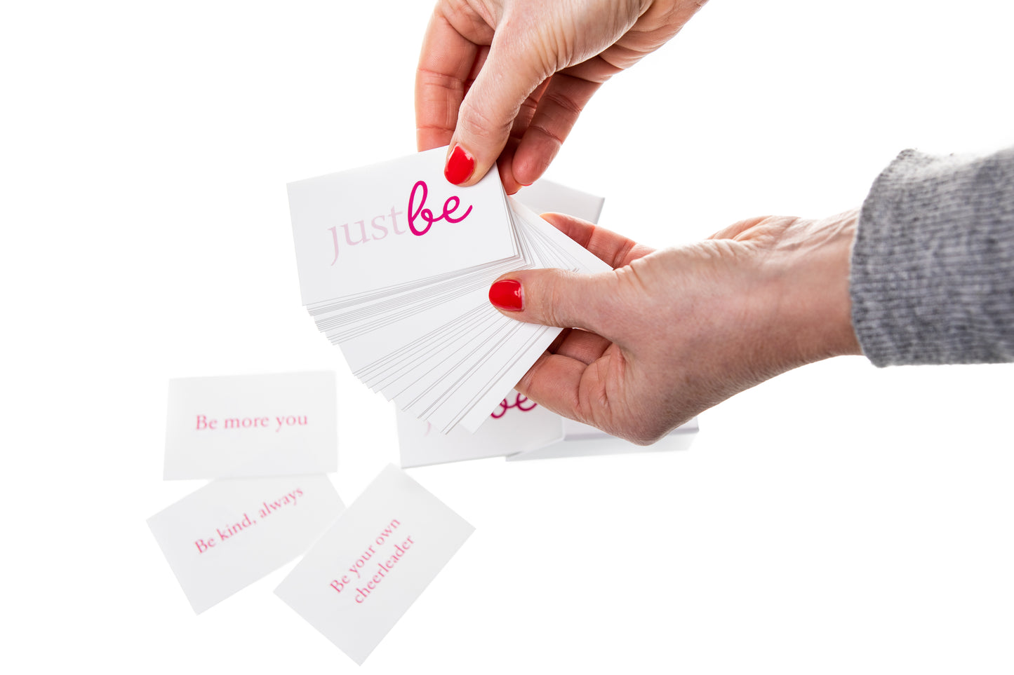 A pair of hands holding JustBe Kind Inspiration Cards with pink text, featuring affirmations like “Be more you” and “Be your own cheerleader” set against a white background.