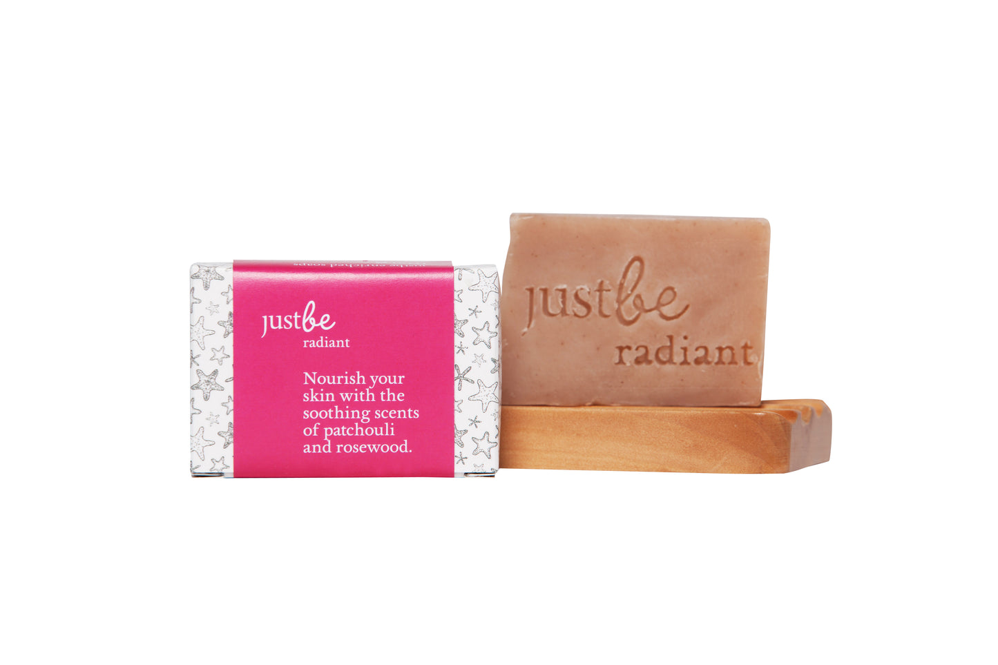 JustBe Radiant Enriched Soap bar with pink and white star-patterned wrapper, displayed next to unwrapped bar on wooden soap dish, embossed with “just be radiant and text "Nourish your skin with the soothing scents of patchouli and rosewood."