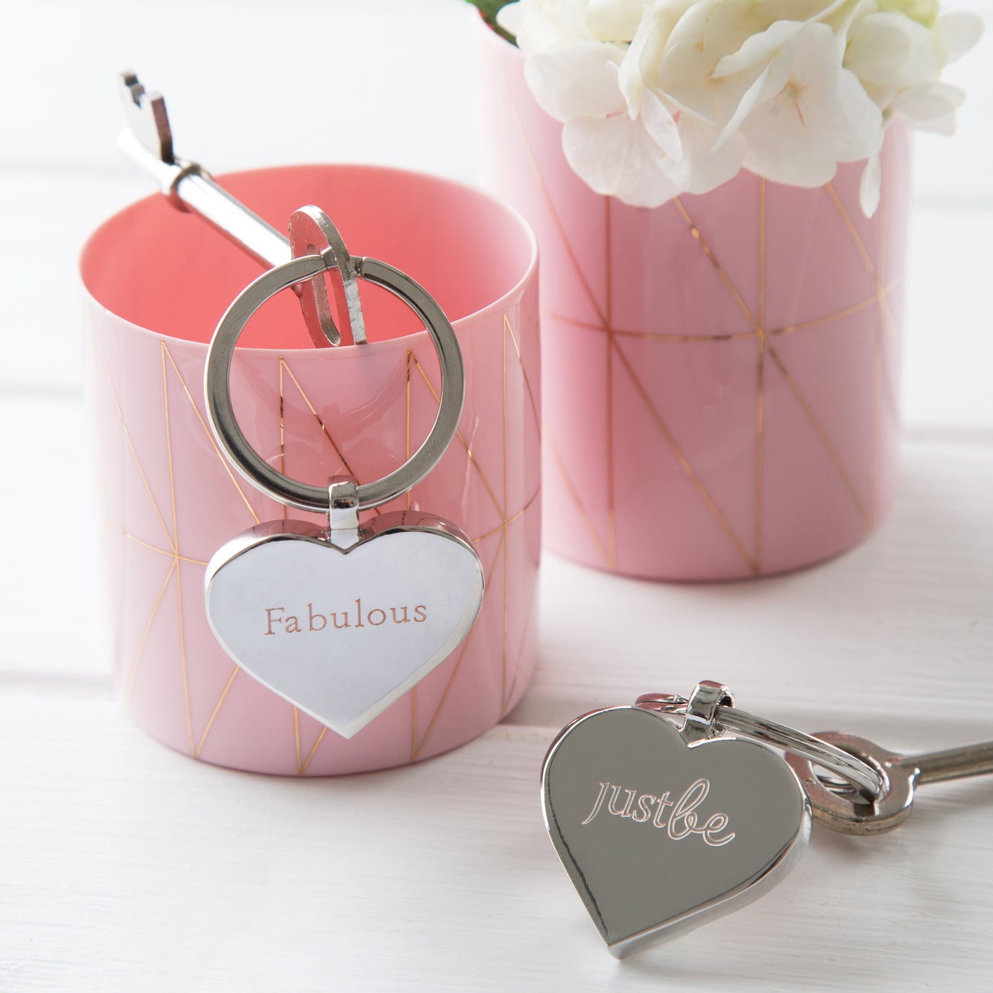 Silver Heart shaped keyring engraved with JustBe and Fabulous on the other. Image shows 2 Keyrings one on top of a pink container, the other sitting on a white surface beside it. White flowers in pink container in the background.