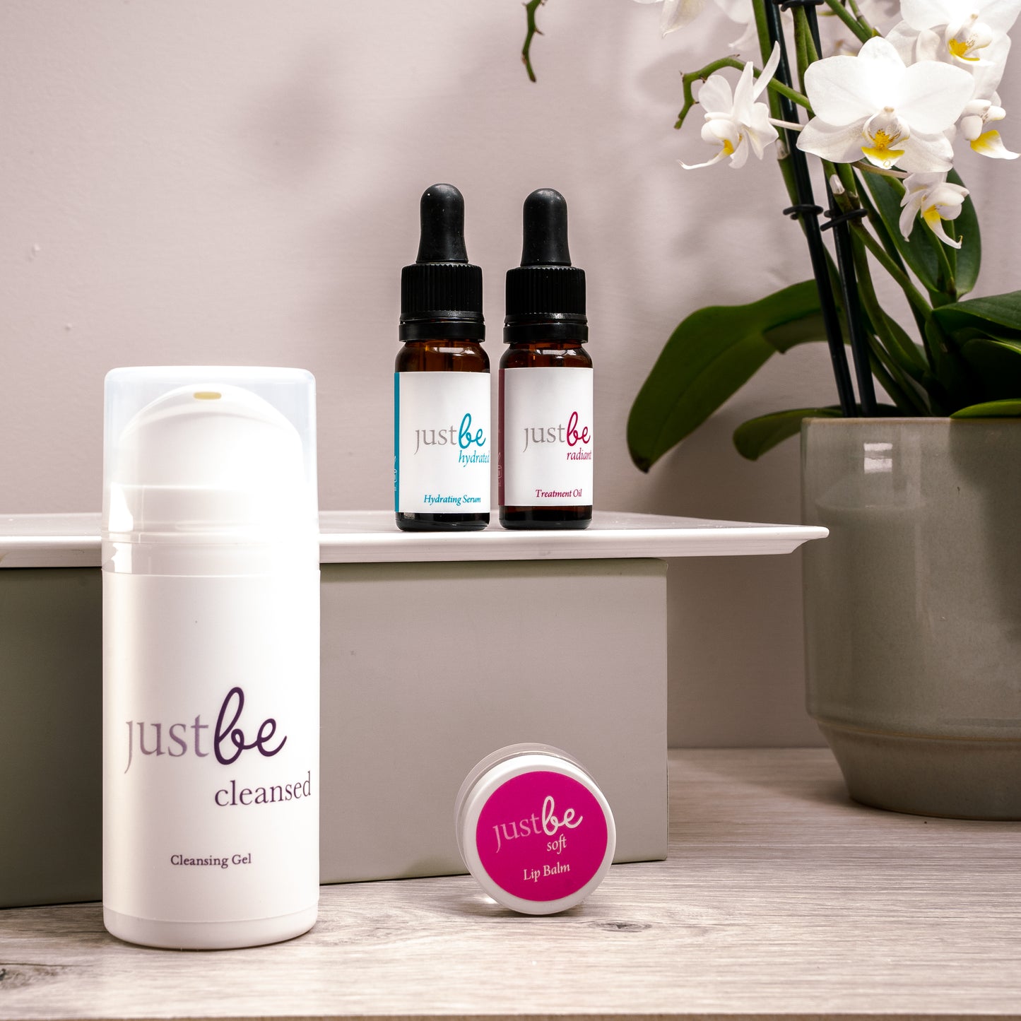 JustBe Botanicals vegan skincare set featuring Cleansing Gel, Hydrating Serum, Treatment Oil and Lip Balm on a wooden surface displayed with white orchid in background