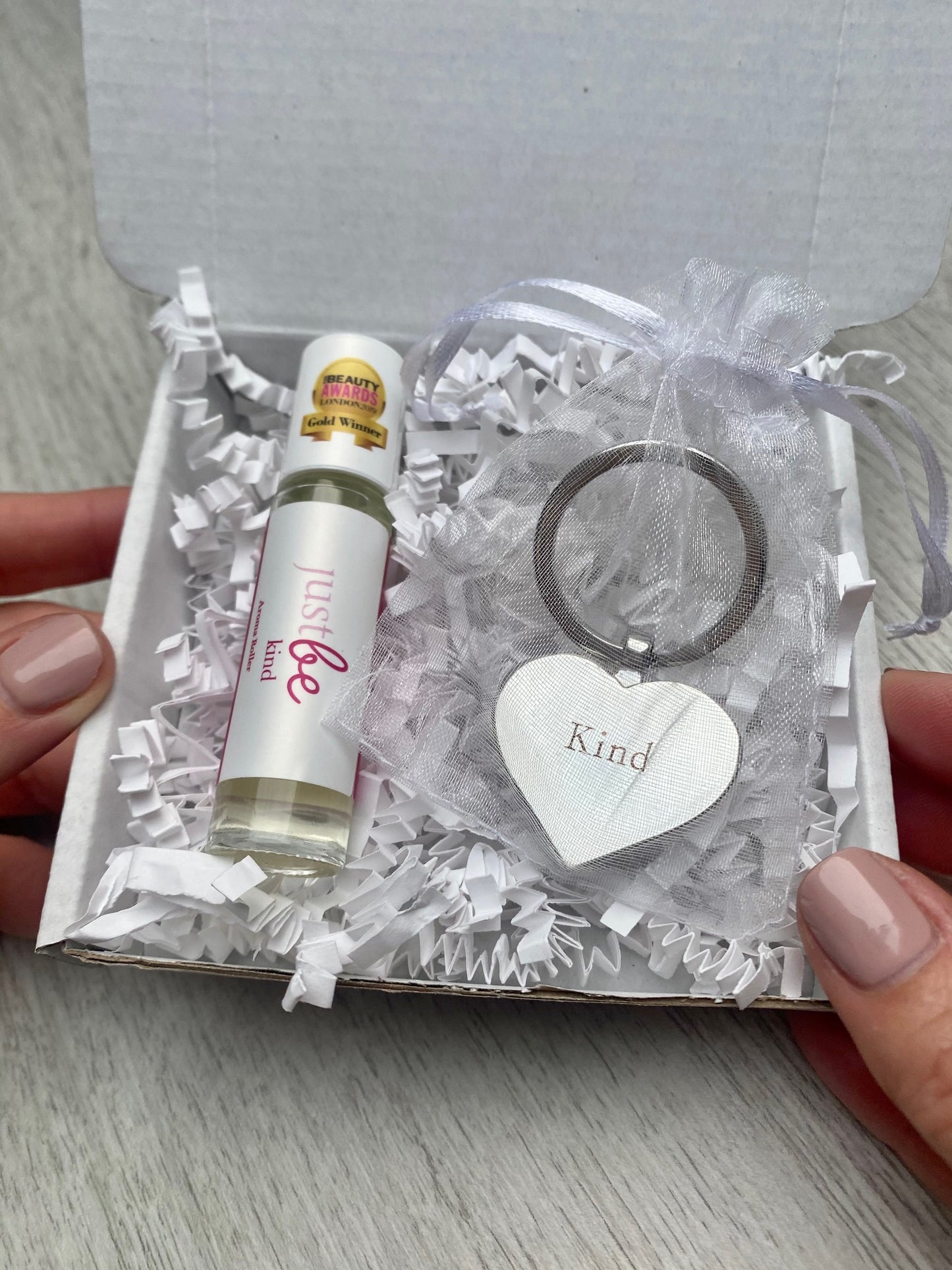 JustBe Kind letterbox gift set with award-winning aromatherapy rollerball and silver heart-shaped keyring engraved with the word kind in organza pouch, presented in white shredded packaging. Being held by hands.