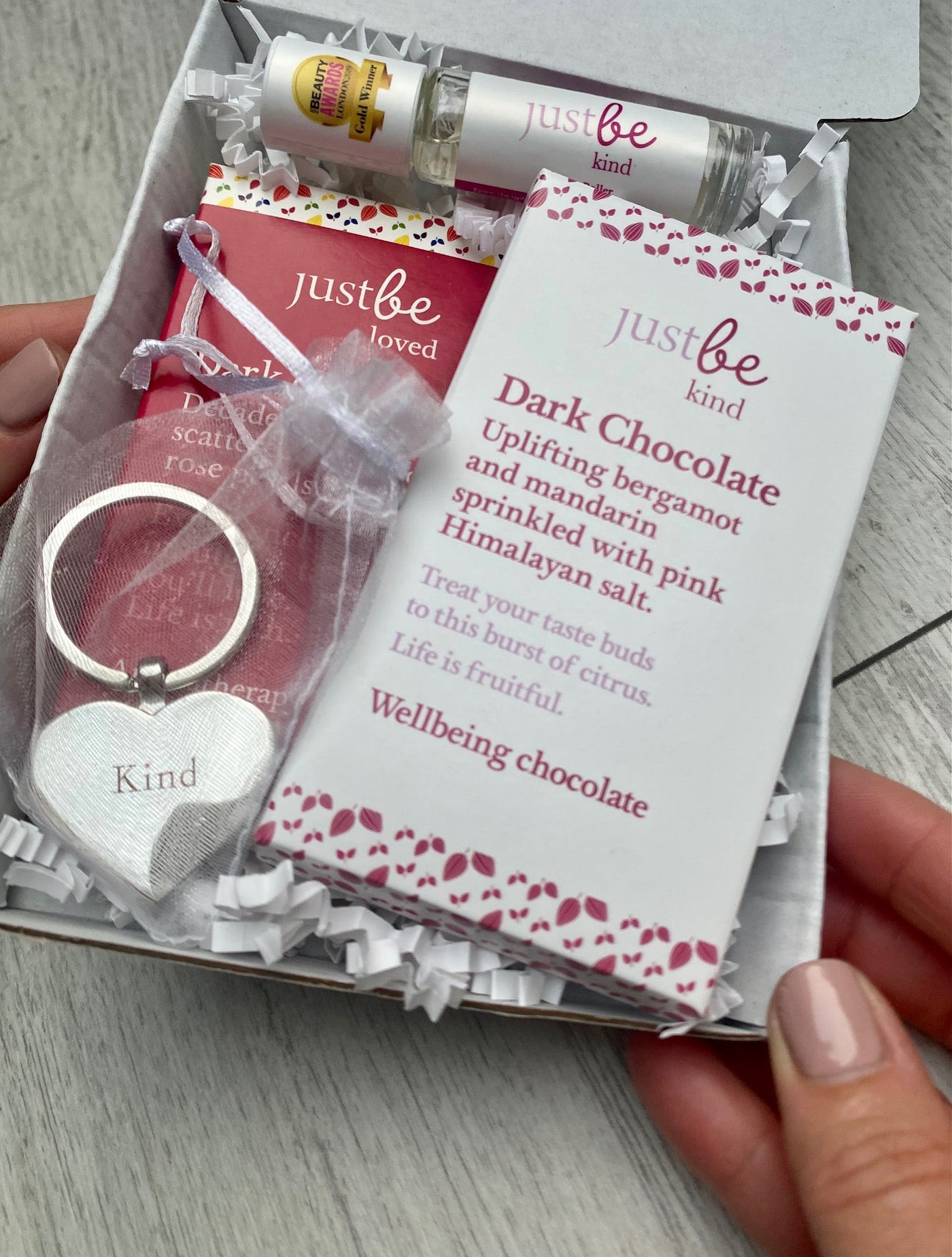 Close-up of a letterbox gift set featuring two JustBe Kind chocolate bars, a heart keyring engraved with the word kind in a white organza pouch, and an aromatherapy rollerball. Box being held.