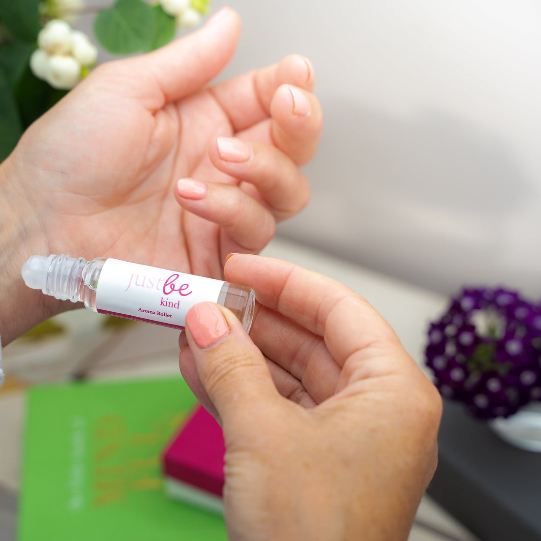Person applying JustBe Kind Aroma Roller to inner wrist, holding a clear glass rollerball bottle with white label and pink text
