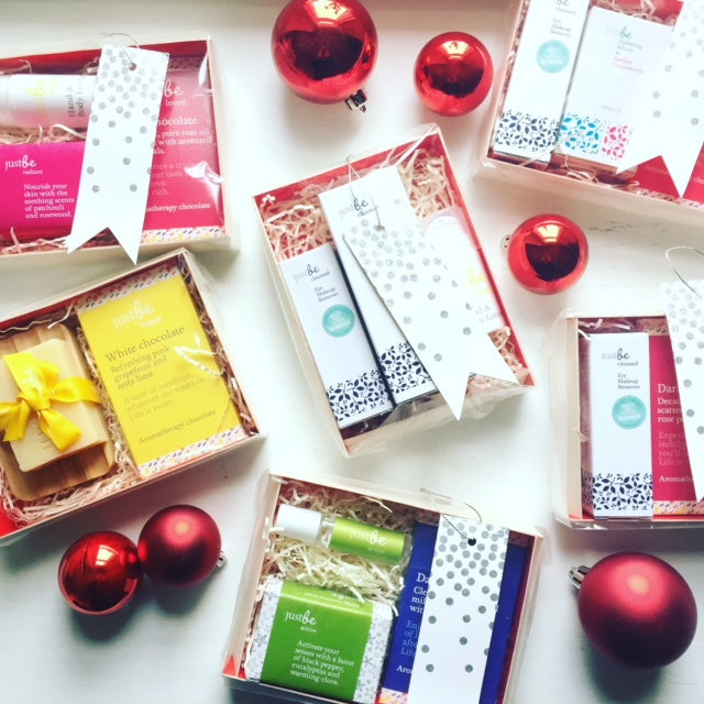 JustBe Botanicals Christmas gift boxes with a mix of chocolate & skincare decorated with white and silver ribbon tags and surrounded by red baubles on a white background