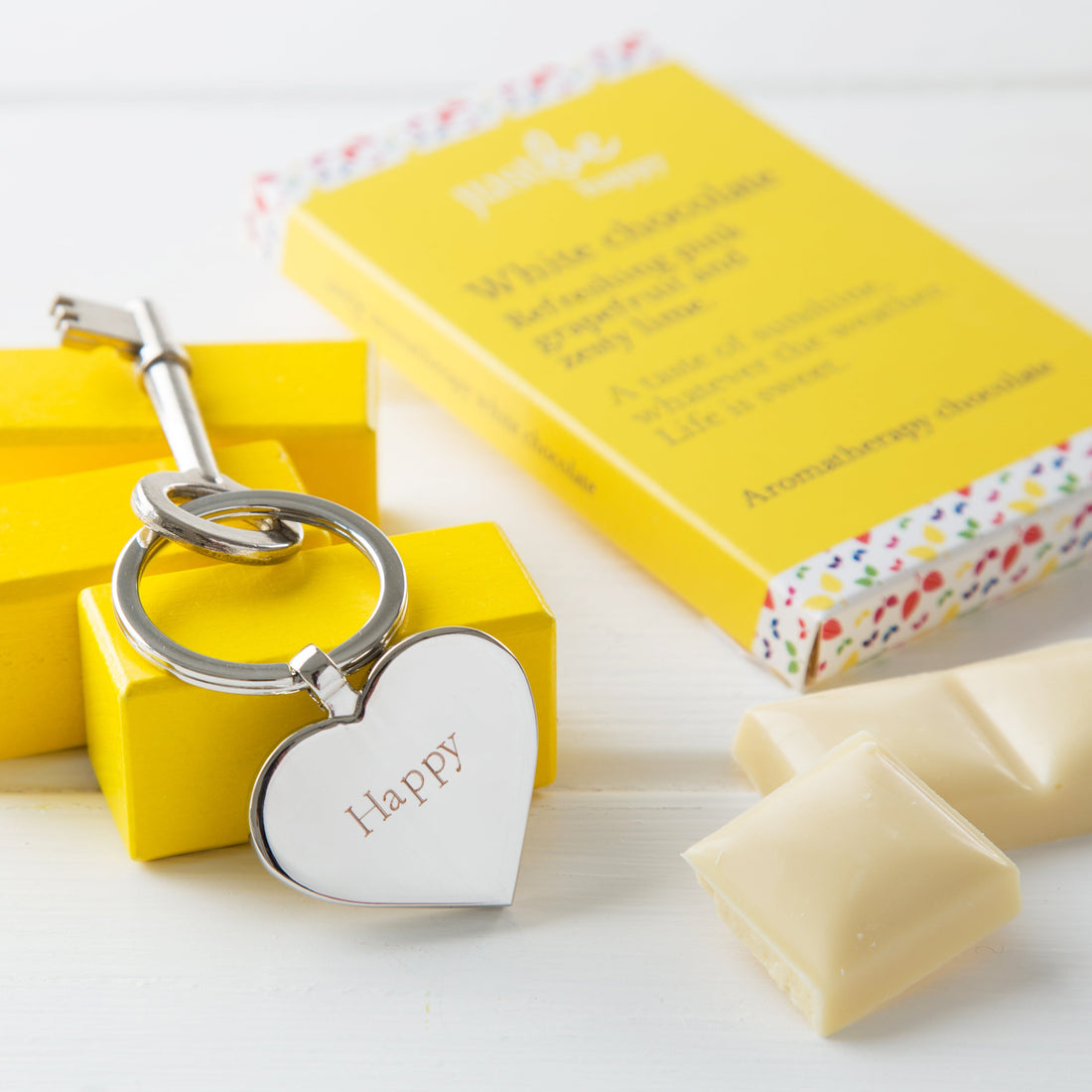 JustBe Happy heart shaped engraved keyring stirring on yellow blocks, sitting beside bar of happy chocolate in bright yellow box