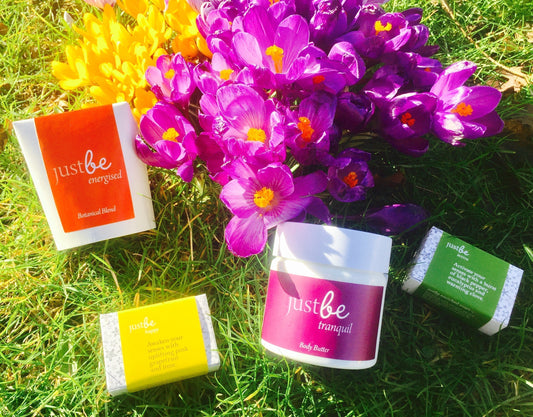 A vibrant spring scene featuring JustBe Botanicals products arranged on grass beside blooming purple and yellow crocuses JustBe Energised herbal tea, JustBe Tranquil body butter, Active and Happy soap bar