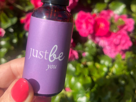 Amber dropper bottle with light purple JustBe You label being held in front of pink rose bush