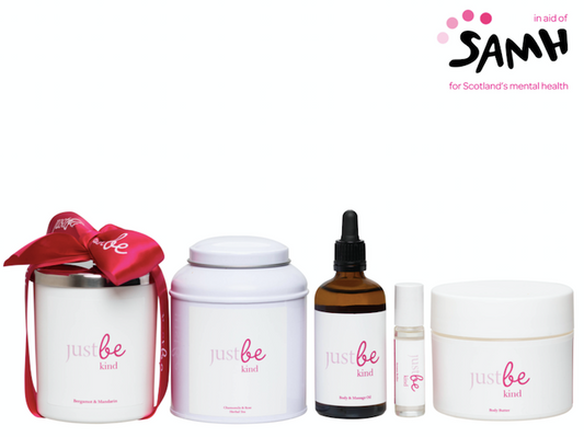 JustBe Botanicals Kind wellbeing range in support of SAMH: five white and pink-labelled products including a candle, tea tin, oil dropper, aroma roller and body butter displayed alongside the SAMH logo.