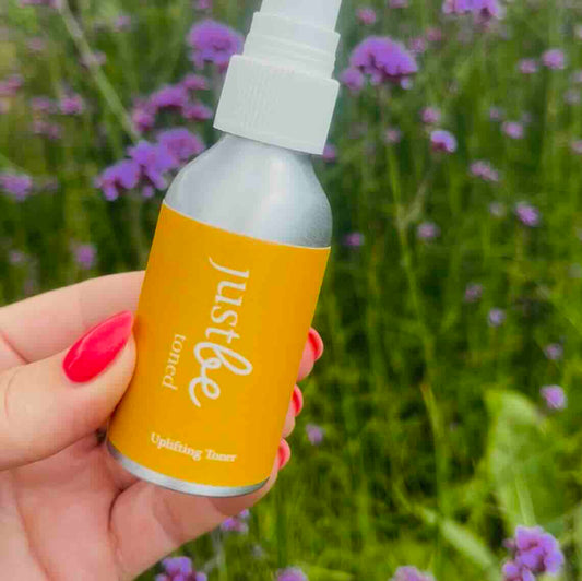 JustBe Botanicals bottle of Uplifting toner with orange label being held against a background of small purple flowers