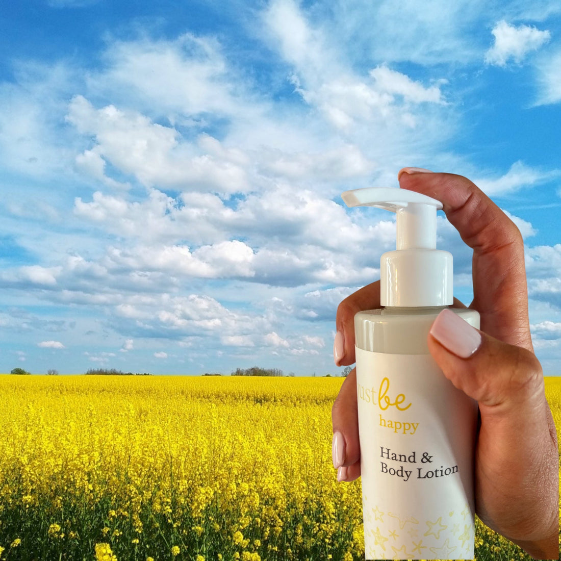 A hand holding a bottle of JustBe Happy Hand & Body Lotion against a bright blue sky and a vibrant yellow flower field, symbolising joy and natural wellbeing