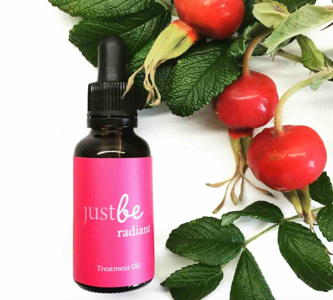 Bottle of JustBe Radiant Treatment Oil sitting beside bright orange rosehips with green leaves