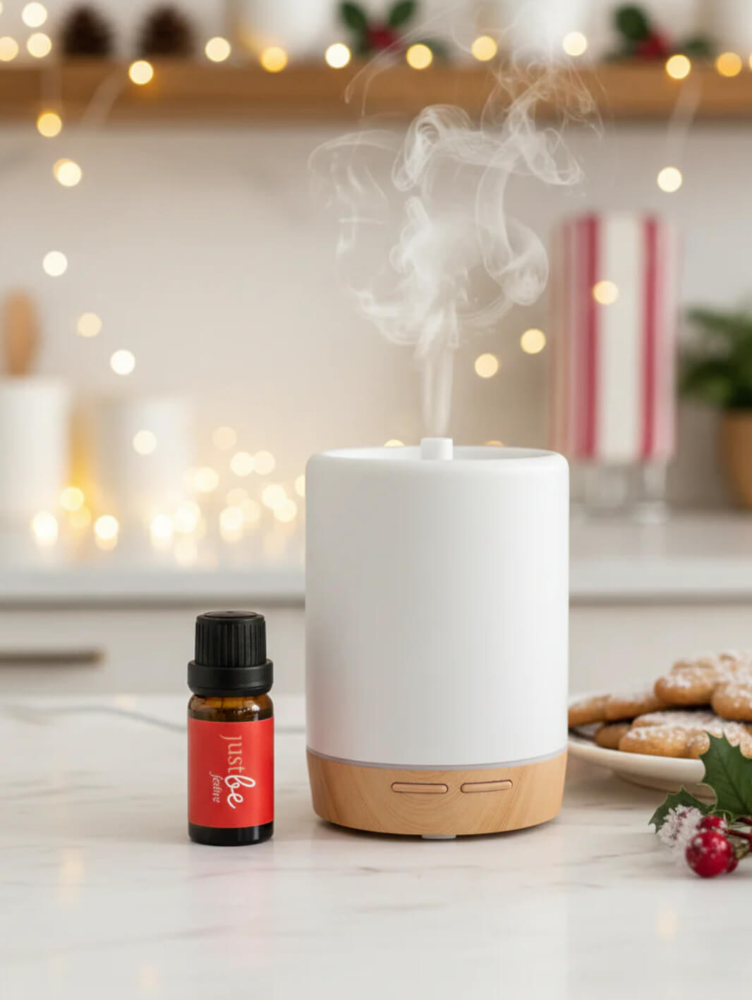 White essential oil diffuser with steam and a bottle of JustBe Festive essential oil on a countertop with festive decorations.