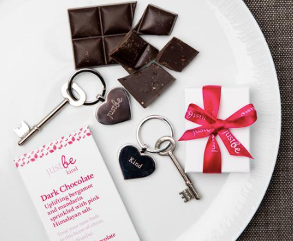 JustBe Kind heart shaped engraved keyring and key on white circular plate along with broken squares of JustBe Kind dark chocolate