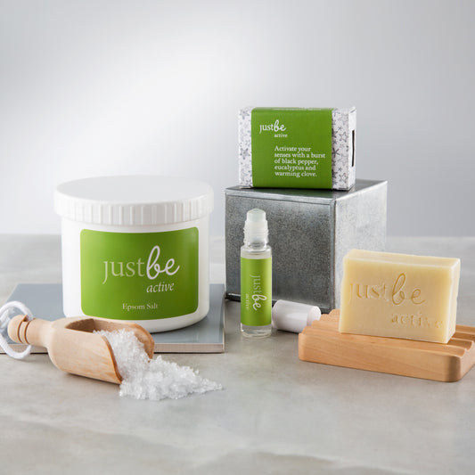 JustBe Active Gift Set featuring Epsom bath salts, aromatherapy rollerball with green label, and enriched soap sitting on top of a wooden rest.  Styled with a wooden scoop with epsom salt spilling out onto a grey surface.