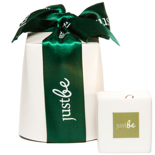 Lit JustBe Active massage candle in a  ceramic square white pot, placed beside a white gift box tied with a dark green JustBe ribbon.