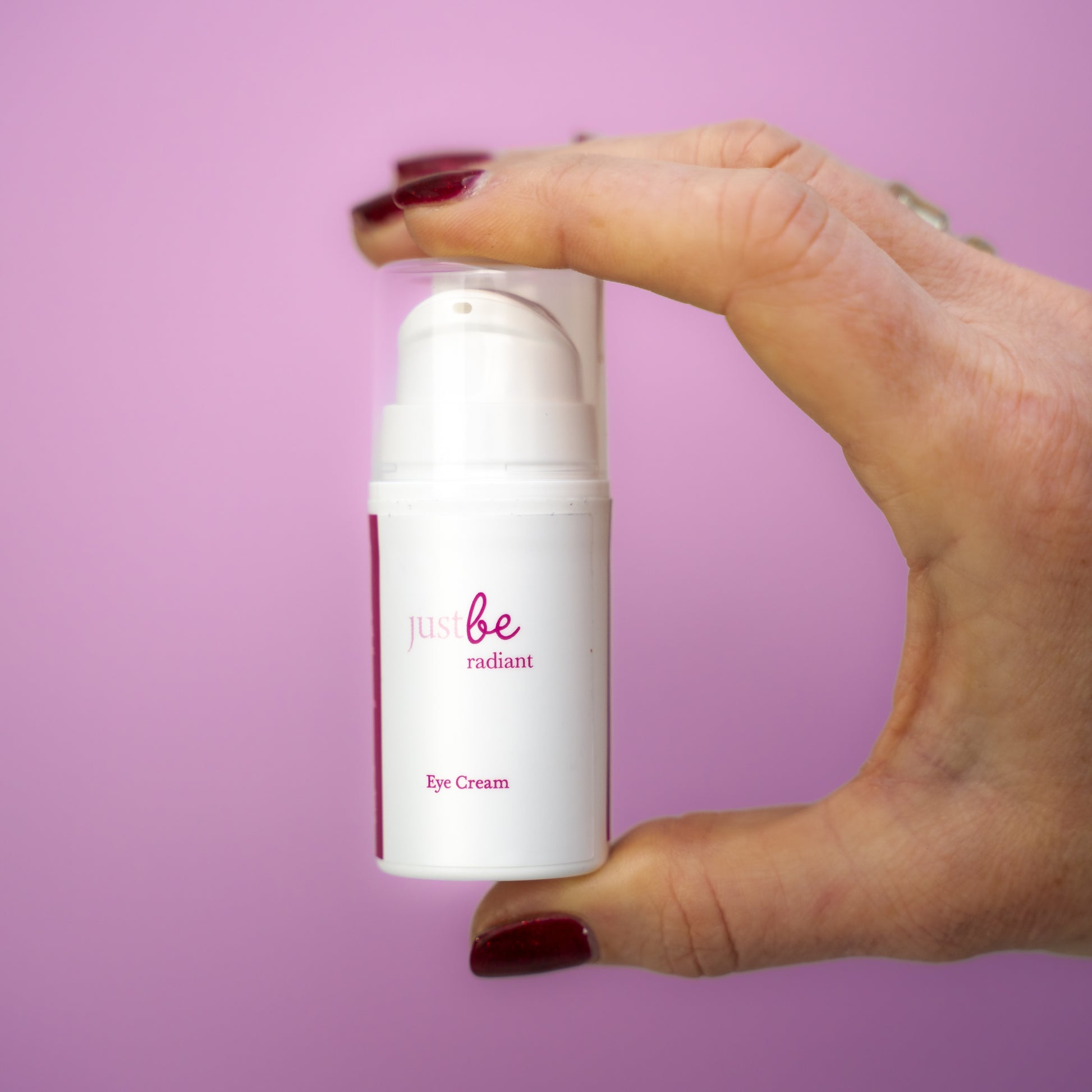 Hand holding JustBe Radiant Eye Cream against a soft pink background, highlighting the compact, travel-friendly packaging