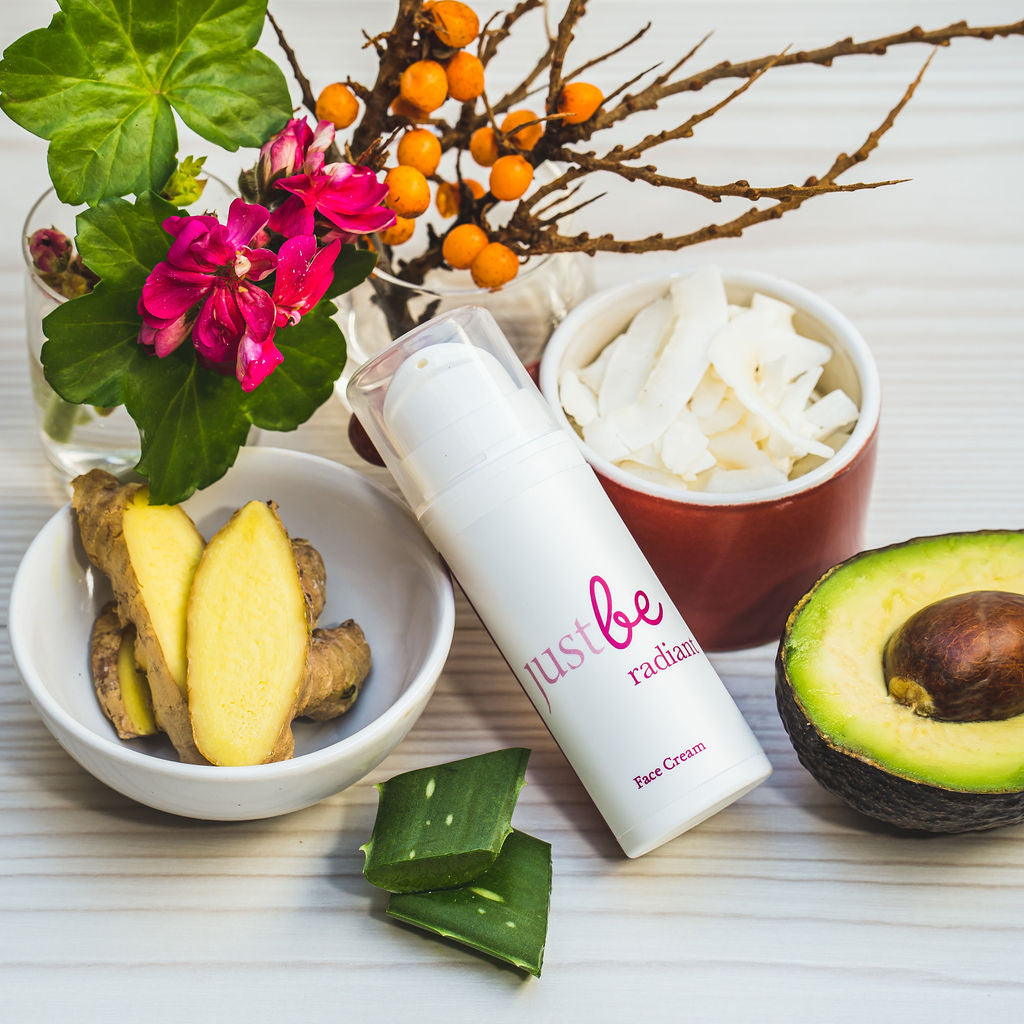 JustBe Radiant Face Cream surrounded by natural ingredients including avocado, aloe vera, fresh ginger, coconut flakes, and sea-buckthorn and geranium flowers  on a light wooden background.