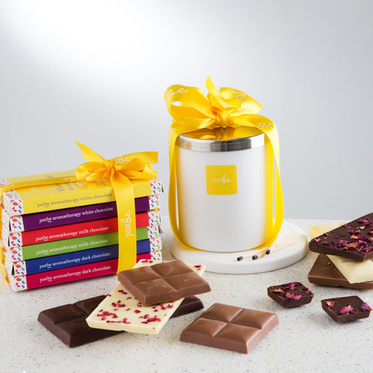 Aromatherapy chocolate gift set with a soy candle in a white container with silver lid wrapped in a yellow ribbon, with 6 chocolate bars tied together with a yellow ribbon. Surrounded by assorted chocolate pieces.