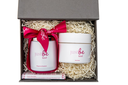 A gift set containing a JustBe Botanicals Kind white aromatherapy candle with a pink label, a jar of body butter, and a dark chocolate bar, all packaged in a box with a pink ribbon.