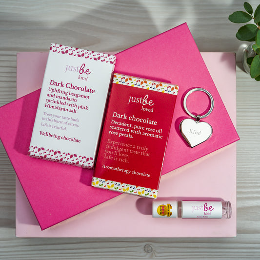 A gift set containing two dark chocolate 50g bars, a heart shaped keyring engraved with 'JustBe' and 'kind' text, and a rollerball with 'JustBe Kind' written on it, all styled on top of pink boxes.