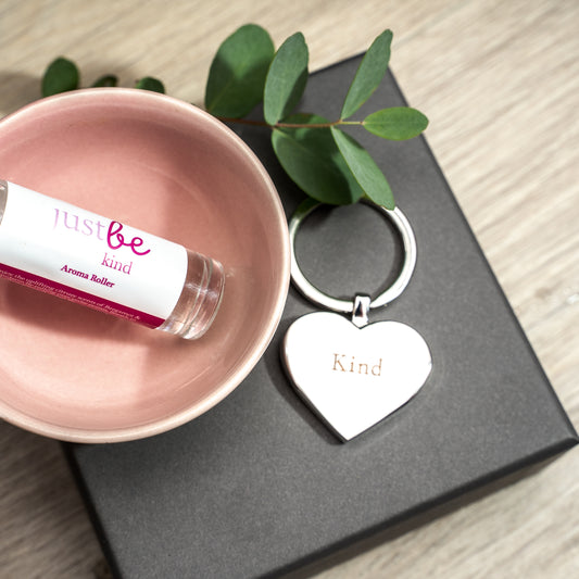 JustBe Kind aromatherapy rollerball in a pink bowl beside a silver heart-shaped keyring engraved with the word kind.  Styled with greenery on a grey gift box.