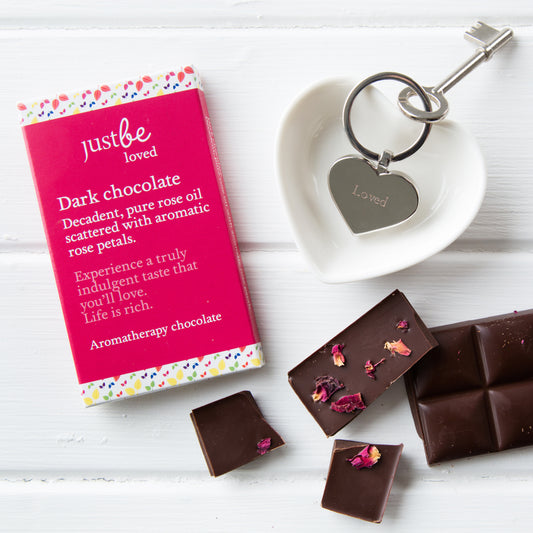 Bar of JustBe Loved dark chocolate styled alongside pieces of the chocolate with rose petals on top and a heart-shaped keyring engraved with the word love.