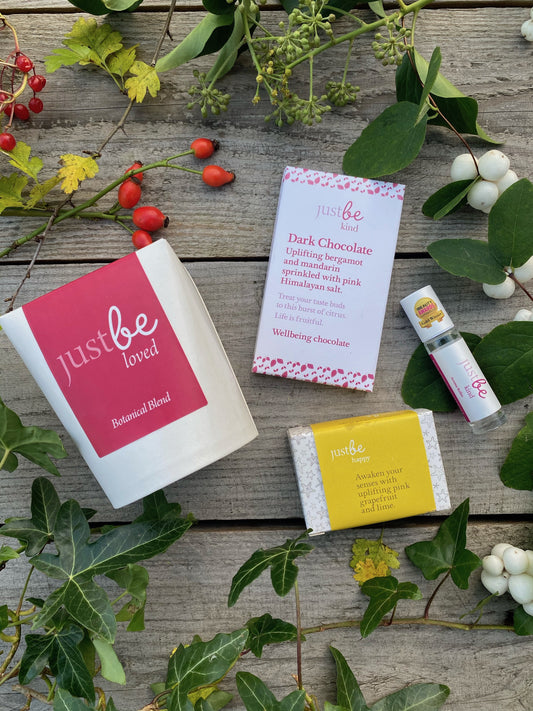 JustBe Botanicals wellbeing gift set including Loved herbal tea, Kind dark chocolate bar, Happy soap bar, and Kind aromatherapy rollerball, styled with greenery on rustic wood.