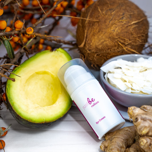 JustBe Radiant Eye Cream surrounded by natural ingredients including avocado, coconut flakes, ginger root and sea buckthorn berries