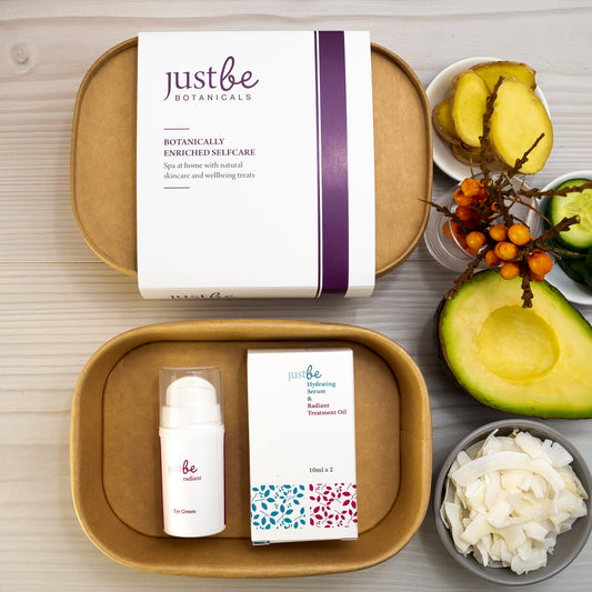 A facial gift set containing JustBe Botanicals skincare products: eye cream in a white pump, and a box containing a glass amber dropper bottles of hydrating serum and treatment oil. Displayed with gift box and beside botanical ingredients: ginger, sea buckthorn, avocado and coconut flakes.