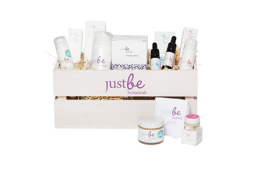 JustBe Botanicals skincare range displayed in white wooden crate with purple JustBe logo on the front. Products displayed inside and also outside crate: eye make up remover, cleansing gel, cleansing balm, hydrating serum, treatment oil, face cream and lip balm arranged with eco-friendly cream straw filler.