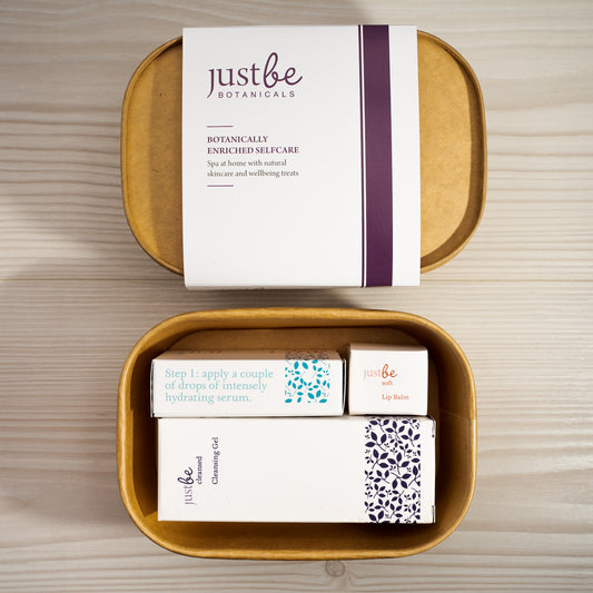 JustBe Botanicals gift box opened to reveal natural skincare products including Cleansing Gel, Hydrating Serum, and Lip Balm presented in eco-friendly packaging on a wooden surface.