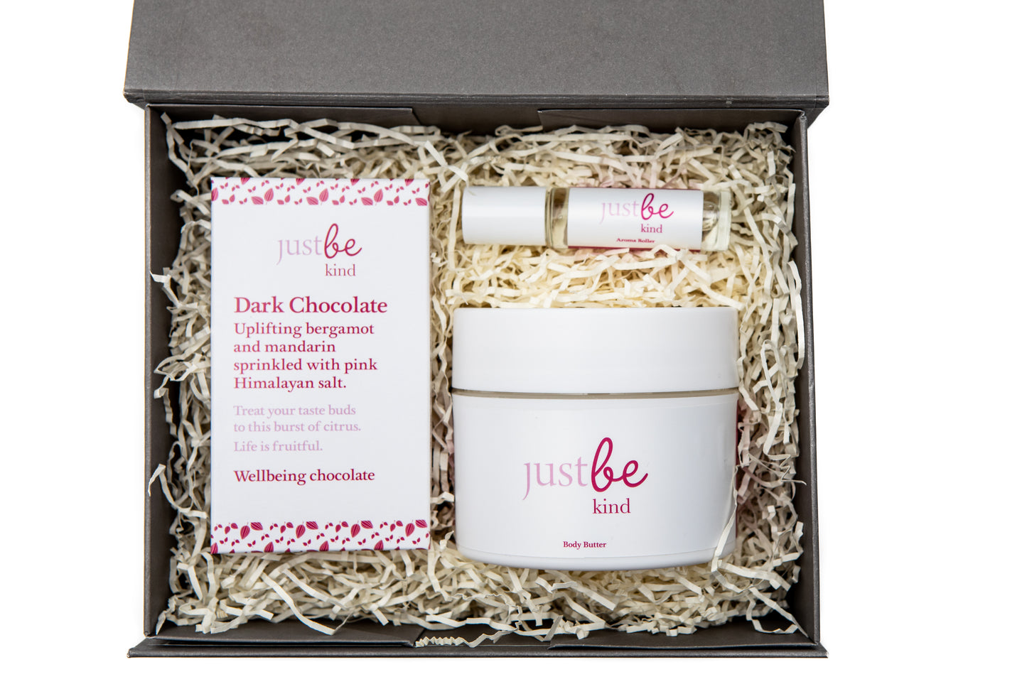 Open grey gift box containing JustBe Kind Body Butter, Kind aromatherapy rollerball, and a bar of Kind Dark Chocolate nestled in shredded paper.