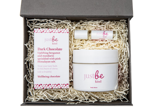 Open grey gift box containing JustBe Kind Body Butter, Kind aromatherapy rollerball, and a bar of Kind Dark Chocolate nestled in shredded paper.
